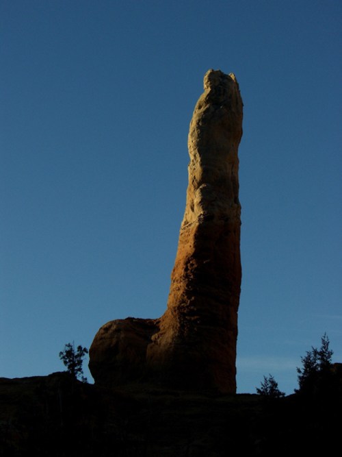 A tall, slender rock formation against a clear blue sky, resembling a unique natural sculpture.
