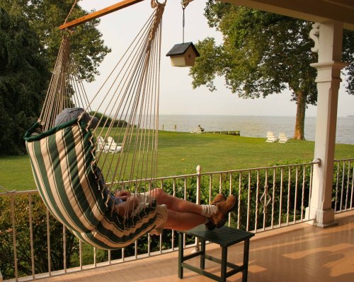 A person relaxes in a striped hammock on a porch, overlooking a serene lake and green lawn.