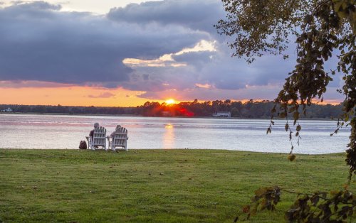 Two people sit on Adirondack chairs by a lake, watching a sunset with colorful clouds reflecting on the water.