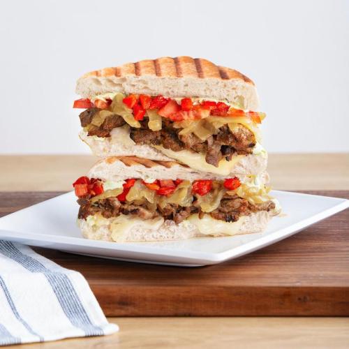 A stacked sandwich with grilled meat, melted cheese, peppers, and onions on toasted bread, served on a white plate.