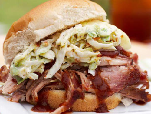 A close-up of a pulled pork sandwich topped with coleslaw and barbecue sauce, served on a soft bun.