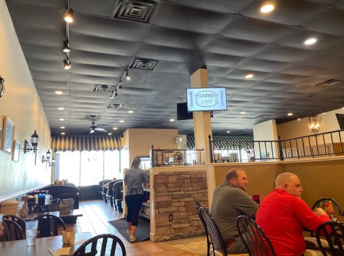 Interior of a cafe with tables, a stone wall, and a TV screen displaying the cafe's name. Two patrons are seated.