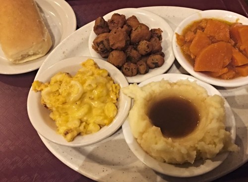 A plate of comfort food featuring macaroni and cheese, mashed potatoes with gravy, fried okra, sweet potatoes, and a roll.