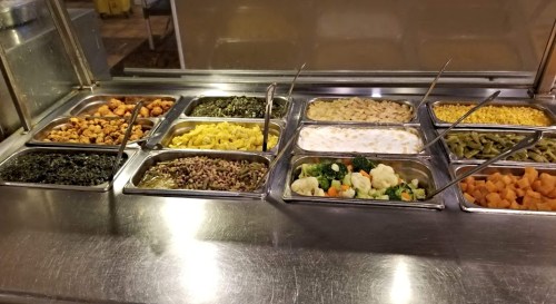 A buffet display with various trays of vegetables, macaroni, and other dishes in a stainless steel serving area.