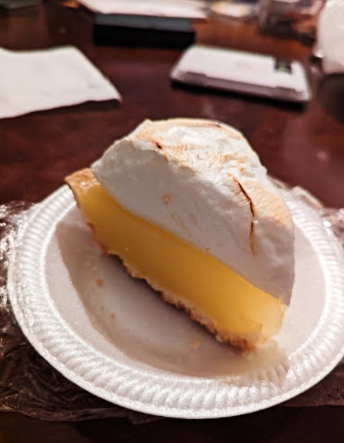 A slice of lemon meringue pie on a paper plate, featuring a smooth yellow filling and a fluffy white meringue topping.