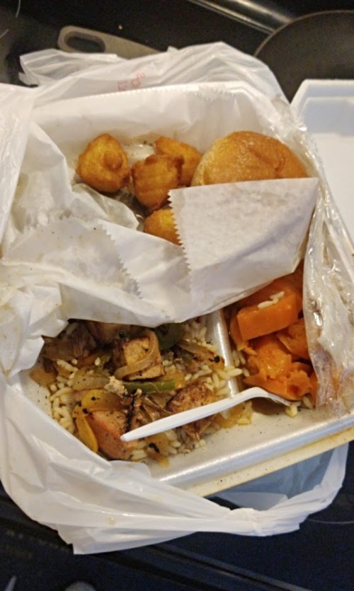 Takeout container with rice, vegetables, sweet potatoes, and fried dough balls, partially wrapped in a plastic bag.