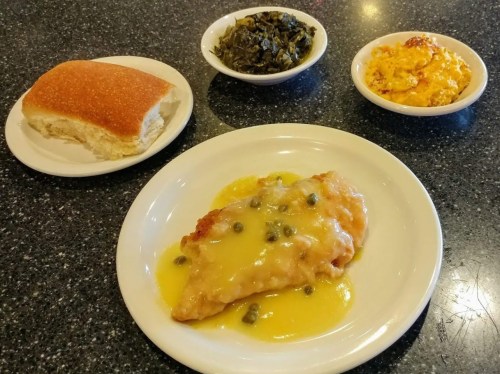 A plate with fish in lemon sauce and capers, served with collard greens, macaroni and cheese, and a bread roll.