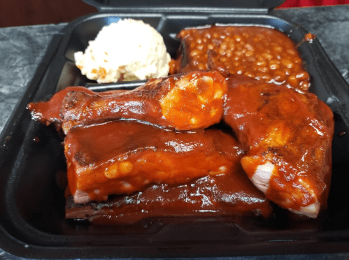 A takeout container with BBQ ribs, baked beans, and coleslaw, all covered in a rich barbecue sauce.