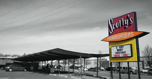 A black-and-white image of Scott's Hamburgers sign with a festive message, featuring a drive-thru area and parked cars.
