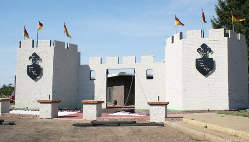 A white castle entrance with two towers, flags on top, and shields on the walls, set against a clear sky.