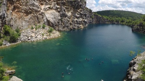 A serene blue lake surrounded by rocky cliffs and lush greenery, with people swimming in the water.