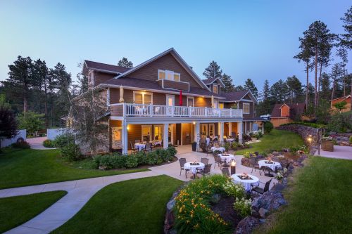 A charming lodge with outdoor seating, surrounded by lush greenery and trees, illuminated at dusk.
