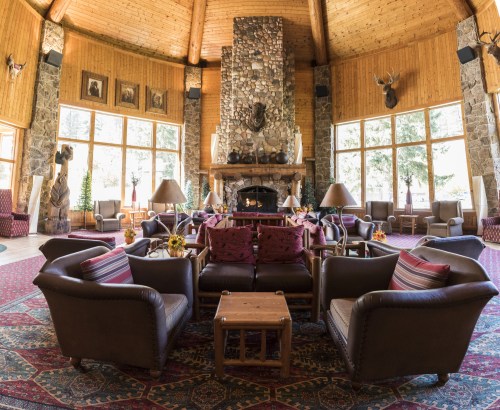 Cozy lodge interior with leather seating, a stone fireplace, and large windows overlooking nature. Warm, inviting atmosphere.
