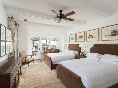 Bright, airy hotel room with two beds, a ceiling fan, and large windows overlooking a balcony. Elegant decor and furnishings.