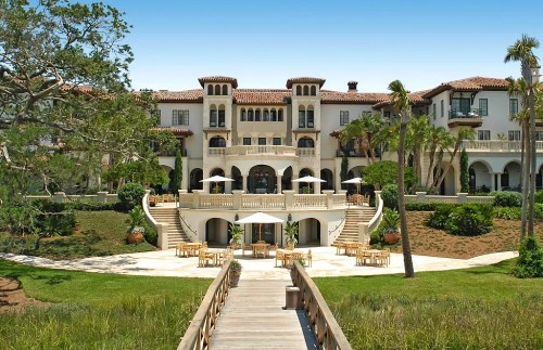 A grand mansion with Mediterranean architecture, surrounded by lush greenery and a wooden walkway leading to outdoor seating.
