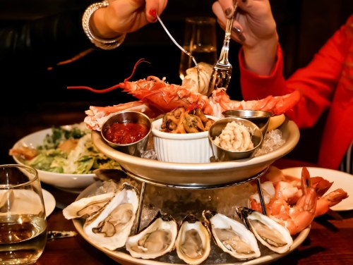 A seafood platter featuring oysters, shrimp, and various sauces, with hands reaching for the food.