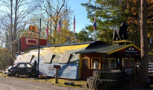 A colorful, rustic building with a gorilla statue on the roof, surrounded by trees and flags, resembling a quirky roadside attraction.