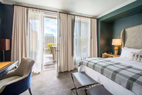 A stylish hotel room with a bed, desk, and large windows opening to a balcony with outdoor seating.