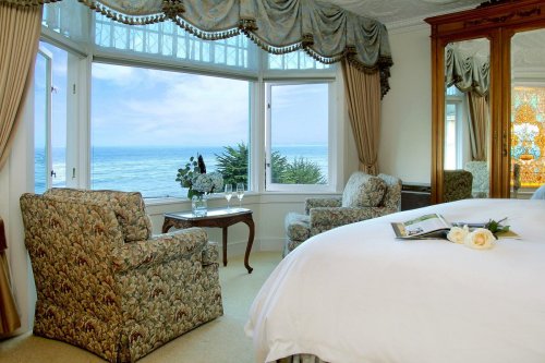 Cozy hotel room with floral chairs, a bed, and a stunning ocean view through large windows.
