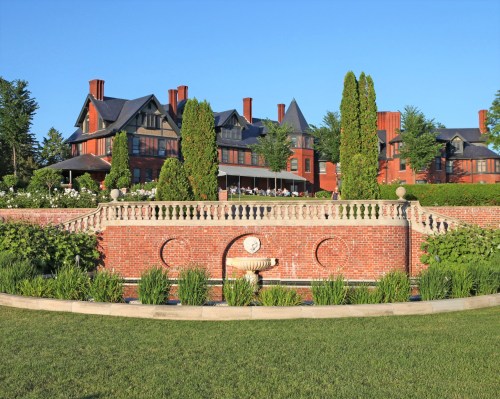 A grand brick mansion with a large lawn and fountain, surrounded by lush greenery under a clear blue sky.