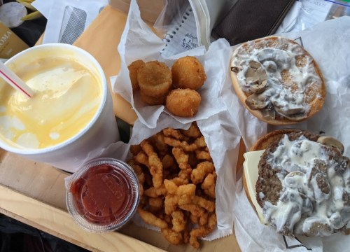 A meal featuring two sandwiches with toppings, a cup of drink, fried snacks, and a small container of sauce.