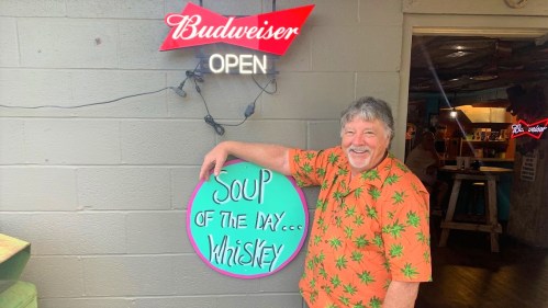 A smiling man in a tropical shirt stands next to a sign that reads "Soup of the Day... Whiskey" under a Budweiser sign.