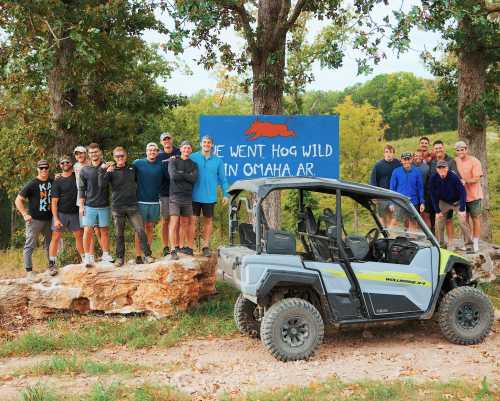 A group of people poses by a sign reading "We Went Hog Wild in Omaha, AR," with an off-road vehicle nearby.