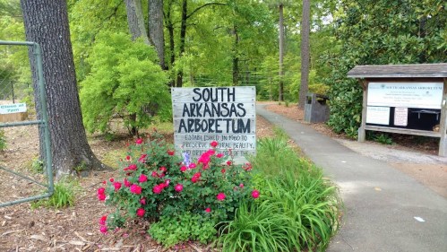 Sign for South Arkansas Arboretum, surrounded by greenery and flowers, with a path leading into the park.