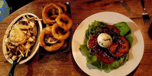 A wooden table with a plate of salad topped with a poached egg, onion rings, and loaded fries in separate dishes.