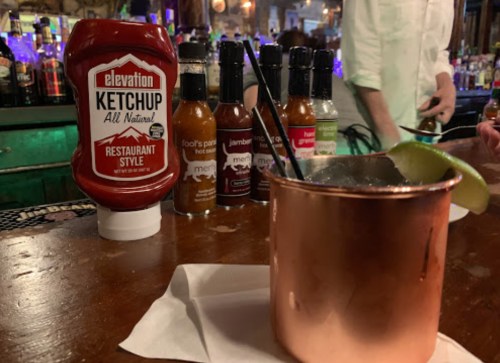 A copper mug with a lime slice sits on a bar, next to a bottle of ketchup and various hot sauces in the background.