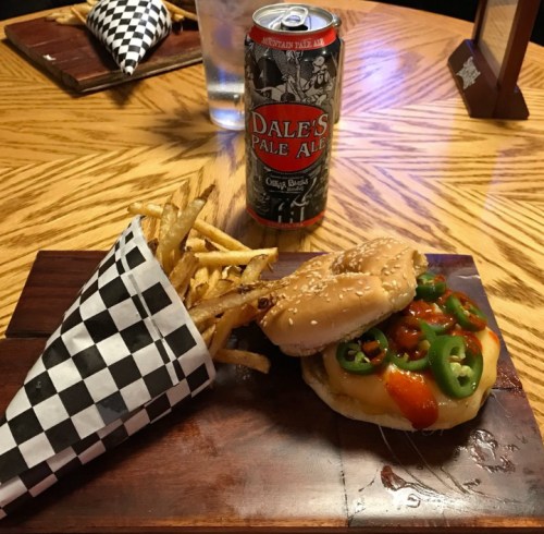 A plate with a burger topped with jalapeños and sauce, served with fries in a checkered cone and a can of pale ale.