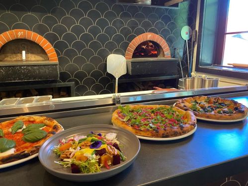 A countertop with four pizzas and a salad, with two brick ovens in the background.