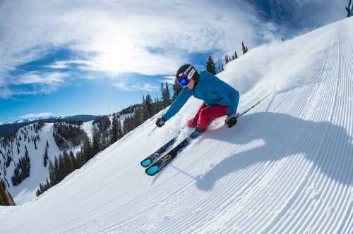 A skier in a blue jacket and red pants carves down a snowy slope under a bright blue sky.