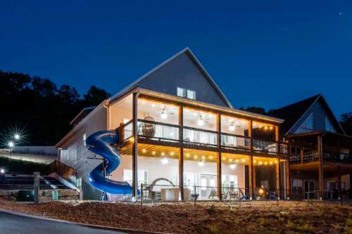 A modern house at night with a blue water slide, illuminated balconies, and surrounding trees.