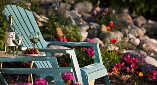 Two turquoise chairs with wine glasses and strawberries, surrounded by colorful flowers and rocks in a garden setting.