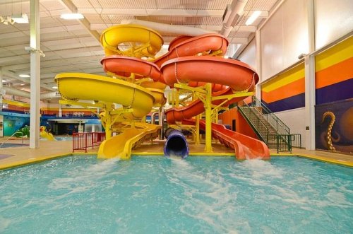 Colorful water slides twist above a pool, with splashes of water and a vibrant indoor water park setting.
