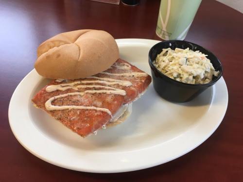 A plate with a salmon sandwich drizzled with sauce and a side of coleslaw in a black bowl.