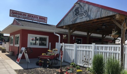 Exterior of Smokin Stan's BBQ, featuring a red building, outdoor seating, and colorful flowers in a garden.