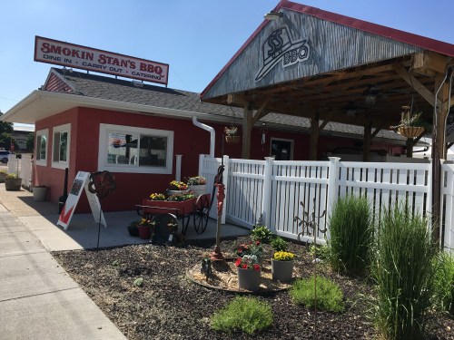 Exterior of Smokin Stan's BBQ, featuring a red building, outdoor seating, and colorful flower beds.