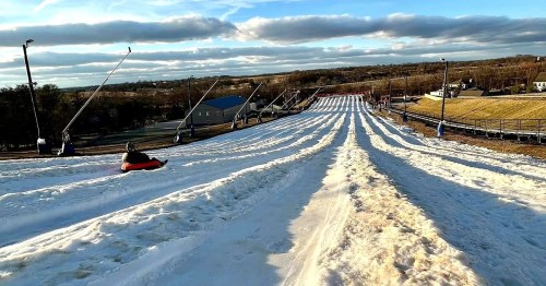 A snowy hill with multiple lanes for tubing, under a cloudy sky, with a person sliding down on a tube.