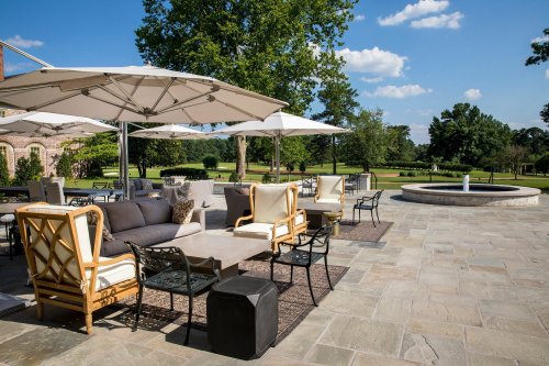 Outdoor patio with comfortable seating, umbrellas, and a fountain, surrounded by lush greenery and blue skies.
