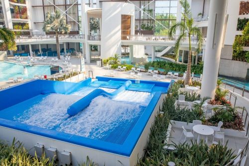 A modern indoor pool area featuring a wave machine, surrounded by palm trees and lounge chairs.