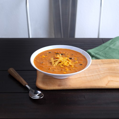 A bowl of creamy soup topped with tortilla strips, placed on a wooden cutting board with a spoon beside it.