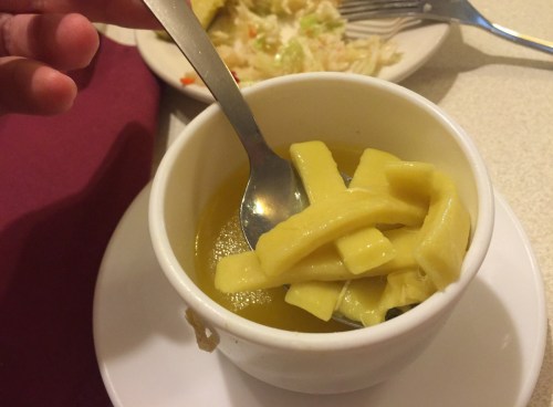 A small bowl of yellow soup with strips of pasta, accompanied by a spoon.