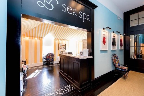 Brightly lit spa reception area with striped walls, a dark wooden desk, and colorful chairs. "Sea Spa" sign above.