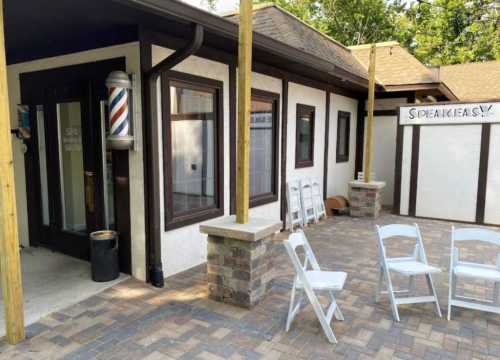 Exterior view of a barbershop with a vintage barber pole and a sign reading "SPEAKEASY," surrounded by chairs.