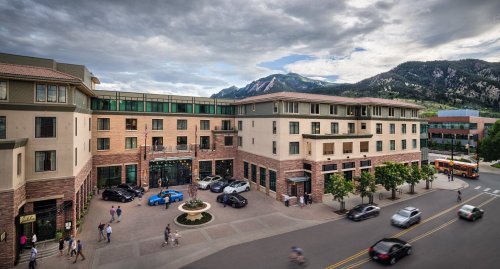 A modern building with shops and cars outside, set against a backdrop of mountains and cloudy skies.