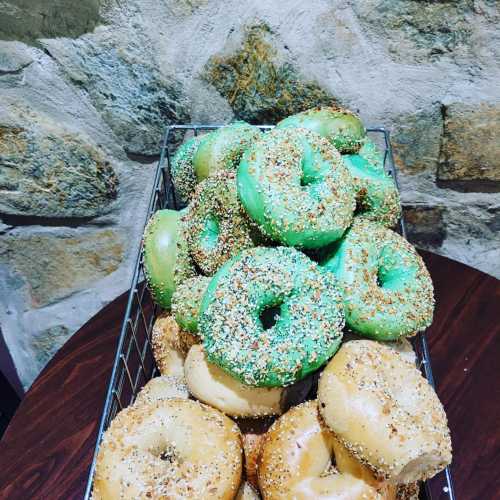 A wire basket filled with green and plain bagels, sprinkled with various toppings, against a stone wall background.