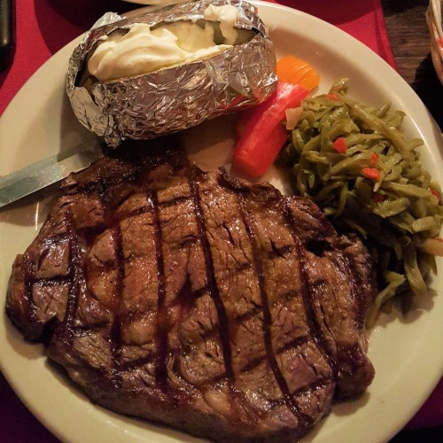 A plate featuring a grilled steak, baked potato with sour cream, and sides of green beans and sliced vegetables.