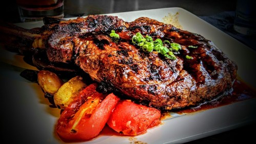 A grilled ribeye steak garnished with green onions, served with roasted vegetables and tomatoes on a white plate.